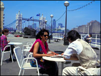 Casal aproveita o dia de sol sentado em um café à margem do rio Tâmisa, em Londres, tendo ao fundo a ponte da Torre de Londres