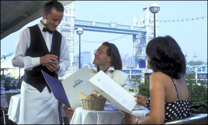 Casal é atendido por um garçom no restaurante Pont de la Tour, à margem do Tâmisa, tendo como pano de fundo a ponte da Torre de Londres